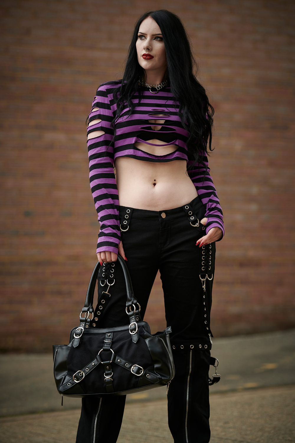 Alternative model in long sleeved crop top with ripped details in black and purple stripe and low rise grunge black trousers 